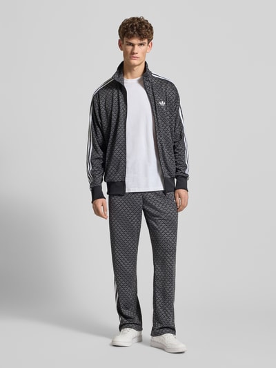 Männermodel trägt Trainingshose mit Logo-Muster von adidas Originals im Relaxed-Fit mit Label-Stitching und elastischem Bund, Ganzkörperansicht von vorne.