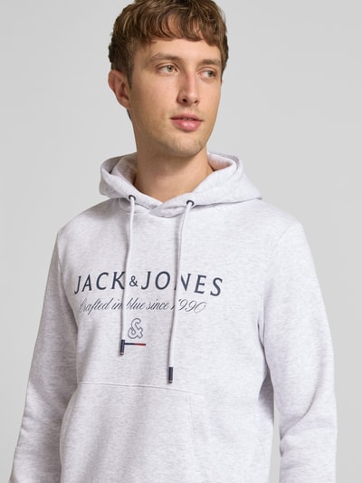 Männermodel trägt Relaxed Fit Hoodie aus katoenmix Model 'KYLE' van JACK & JONES in lichtgrijs met labeldetail en Kangoeroezak, Nahaufnahme Oberkörper. Ruglengte maat M: 67 cm, mouwlengte maat M: 65 cm.