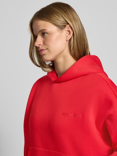 Frauenmodel trägt Kapuzenpullover aus Baumwoll-Mix von REVIEW in leuchtendem Rot mit Känguru-Tasche, Nahaufnahme Oberkörper.