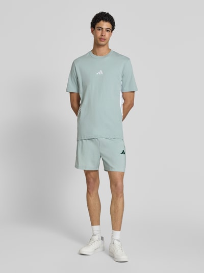 Męski model nosi Regular Fit Sport-Shorts mit Kontraststreifen Modell 'CHELSEA' od adidas Sportswear w jednolitym jasnoniebieskim kolorze, z logo, elastycznym paskiem i bocznymi kieszeniami, całopostaciowy widok z przodu.