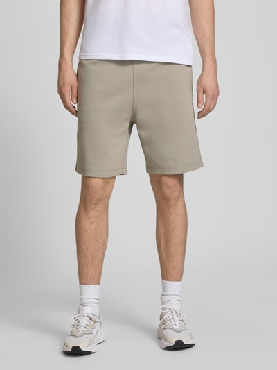 Mężczyznamodel nosi Shorts mit Label-Stitching mit elastischem Bund von CHAMPION, beżowe, bawełniany miks, krój regular fit, szerokość w rozmiarze M 37 cm, długość nogawki 42 cm, widok z przodu na całe ciało.