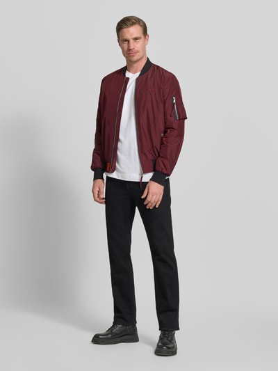 Męski model nosi Bomberjacke mit Label-Patch und Eingrifftaschen od Original Bombers, prosty krój, materiał bordowy z czarnymi ściągaczami, długość tyłu w rozmiarze M: 67 cm, szerokość w rozmiarze M: 53 cm, szerokość ramion w rozmiarze M: 43 cm, długość rękawa w rozmiarze M: 68 cm, widok całościowy z przodu.