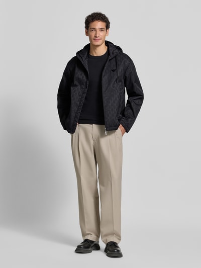 Männermodel trägt Regular Fit Jacke mit Allover-Logo-Print von Emporio Armani in Schwarz mit Kapuze und Eingrifftaschen, Ganzkörperansicht von vorne.