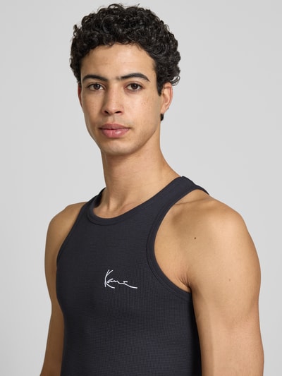 Mannelijk model draagt Tanktop met Label-Stitching van KARL KANI in zwart katoenmix met structuurpatroon, slim fit, ronde hals. Nahaufnahme Oberkörper.