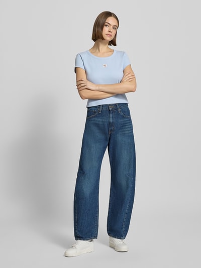 Frauenmodel trägt Cropped T-Shirt mit Label-Detail von Calvin Klein Jeans z mieszanki bawełny i elastanu, krój krótki, okrągły dekolt, długość tyłu 43 cm dla rozmiaru S, jasnoniebieski, prosta tekstura, jeansy, białe buty, całkowity widok z przodu.