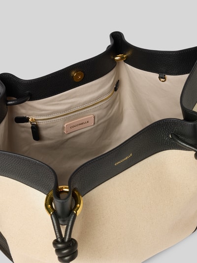 Produktfoto, Shopper mit Label-Detail Modell 'FINN CANVAS' von Coccinelle, beige-schwarze Tasche aus Canvas und Leder, goldene Details, Innenansicht mit Reißverschlussfach, Druckknopfverschluss, lange Tragegriffe, Maße 60x34x17 cm, Schulterriemen 74 cm.