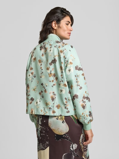 Vrouwenmodel draagt Blouson im Allover-Look mit Stehkragen van Marc Cain in mintgroen met bruine bloemmotieven, regular fit, knoopsluiting, klepzak, ruglengte maat 36: 54 cm, schouderbreedte: 43 cm, borstwijdte: 58 cm, mouwlengte: 64 cm, all-over look, ganzlichaamsaanzicht achterzijde.