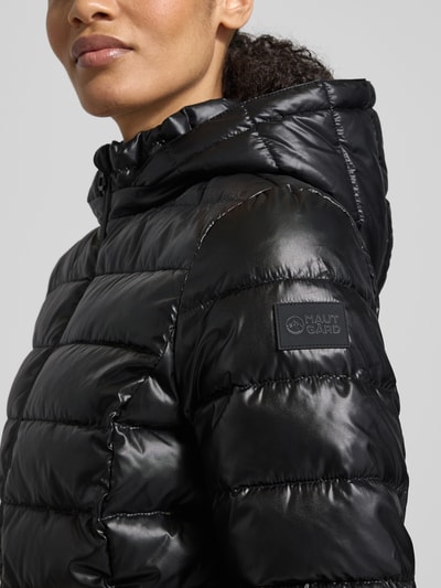 Frauenmodel trägt Jacke mit Kapuze von NAUTGÅRD in Schwarz mit Label-Detail, wattierte Textur, Nahaufnahme Oberkörper. Lockere Passform.