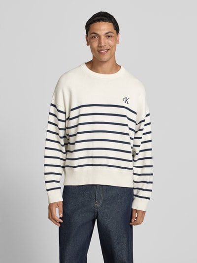 Männermodel trägt Regular Fit Strickpullover aus reiner Baumwolle van Calvin Klein Jeans in gebreide look met gestreept motief, logostitching, ronde hals en lange mouwen, puur katoen, kleuren crème en marineblauw, geribde boorden, Ganzkörperansicht von vorne.