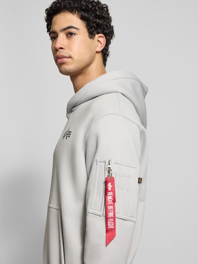 Männermodel trägt Hoodie mit Label-Details von Alpha Industries, Baumwoll-Mix, Lockerer-Schnitt, Kapuze, Reißverschlusstasche am Ärmel, Känguru-Tasche, gerippte Abschlüsse, Rückenlänge 68 cm, Ärmellänge 67 cm, Nahaufnahme Oberkörper.