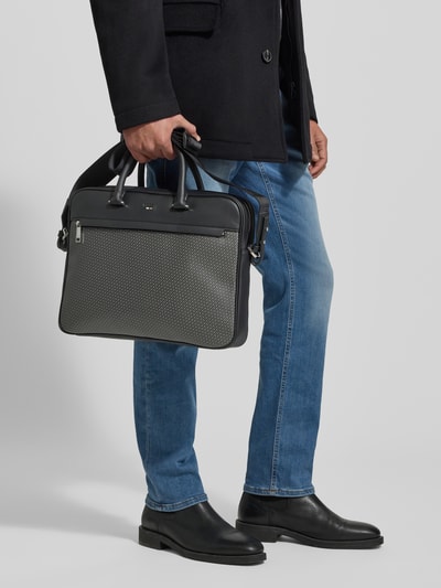 Männermodel trägt Laptoptasche mit Monogramm Muster Modell 'RAY_MN_S' von BOSS in Schwarz mit Tragehenkel, Ganzkörperansicht von vorne.