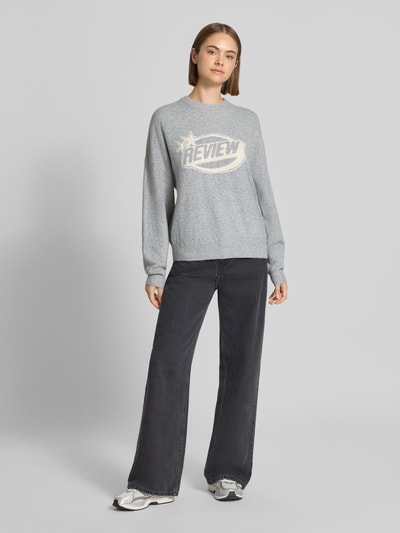 Kobiece model nosi sweter z dzianiny 'Strickpullover mit Label-Print' od REVIEW, luźny krój, okrągły dekolt, nadruk z logo, szeroko opadające ramiona, prążkowany dół, długość tyłu w rozmiarze S: 56 cm, długość rękawa w rozmiarze S: 44 cm; widok całej sylwetki z przodu.