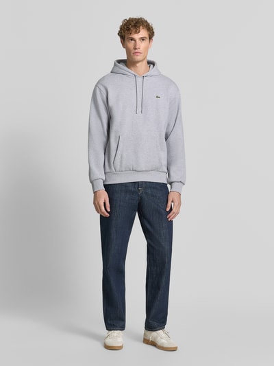 Męski model nosi Relaxed Fit Hoodie aus Baumwoll-Mix od Lacoste, jasnoszary, kieszeń z przodu, bluza, jeansy, sneakersy, Ganzkörperansicht von vorne, Herren Sweatshirt von Lacoste.