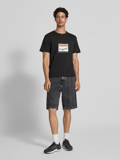Męski model nosi t-shirt z nadrukiem z napisem model 'RHYERES' od Jack & Jones, czarny, krój regular fit, woskowana bawełna, okrągły dekolt, długość tyłu w rozmiarze M: 67 cm, widok z przodu, cała sylwetka