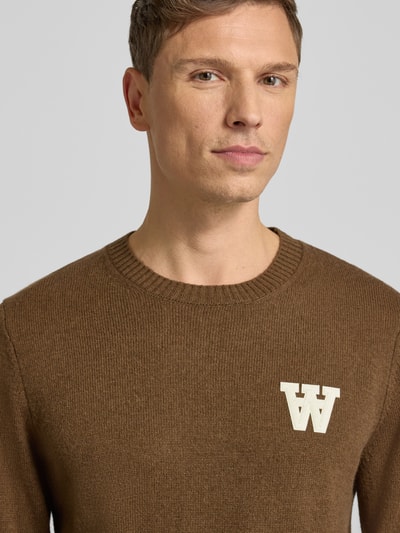 Männermodel trägt braunen Strickpullover aus Lammwolle Modell 'Tay' von Wood Wood mit Label-Stitching, Rundhalsausschnitt, Nahaufnahme Oberkörper.