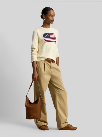 Frauenmodel trägt Strickpullover mit Flaggen-Motiv und Rundhalsausschnitt von Polo Ralph Lauren, Cremefarbe, kombiniert mit beigefarbener Stoffhose, Nahaufnahme Oberkörper. Regular Fit, reine Baumwolle.
