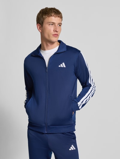 Männermodel trägt Trainingsanzug mit elastischen Abschlüssen von adidas Sportswear in Dunkelblau, Label-Details, prosta krój, Ganzkörperansicht von vorne, Szerokość w rozmiarze M: 36 cm, Długość tyłu: 71 cm, Szerokość ramion: 46 cm, Długość rękawów: 63 cm, Długość wewnętrzna nogawki: 75 cm, wpuszczane kieszenie, elastyczny pas.