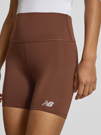Frauenmodel trägt High Rise Radlershorts mit Logo-Print Modell 'HARMONY' von New Balance, braun, elastischer Bund, Nahaufnahme Oberkörper.