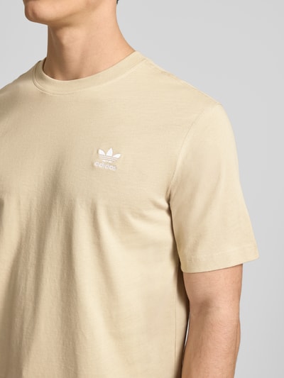 Männermodel trägt T-Shirt mit Label-Stitching von adidas Originals in Beige, reine Baumwolle, Regular Fit, Nahaufnahme Oberkörper.