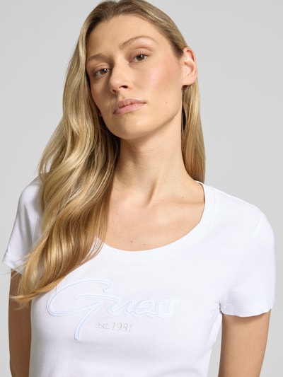 Frauenmodel trägt Damen T-Shirt mit U-Ausschnitt Modell 'SCRIPT' von Guess, weiß, Baumwoll-Elasthan-Mix, körpernaher Schnitt, Label-Stitching, Nahaufnahme Oberkörper.