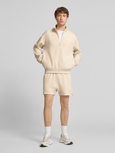 Männermodel trägt Regular Fit Sweatjacke mit Baumwoll-Anteil von adidas Sportswear, Optik: Unifarben, Passform: Regular-Fit, Verschluss: Reißverschluss, Applikation: Logo-Print, Kragenform: Stehkragen, Ärmelform: Langarm, hellbeige Farbe, glatte Struktur, Ganzkörperansicht von vorne.