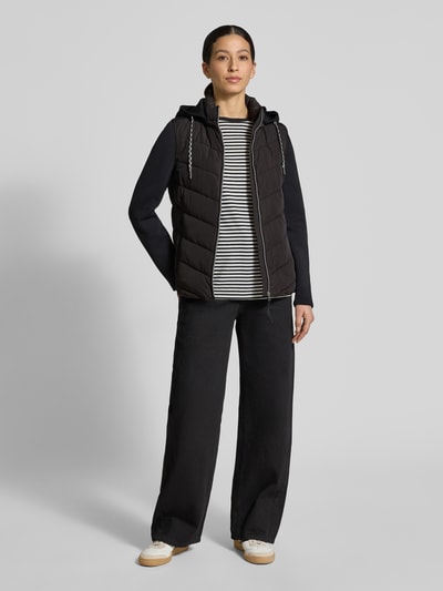 Frauenmodel trägt schwarze Slim Fit Steppjacke mit gefüttertem Stehkragen von Christian Berg Woman Selection, seitlich weiß-schwarzes Streifenshirt und weite Hose, Stepp-Optik, Sicht auf Kapuze und Reißverschlusstaschen, Ganzkörperansicht von vorne.
