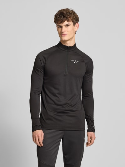 Männermodel trägt PUMA x HYROX - Regular Fit Longsleeve mit Label-Print in Schwarz mit Stehkragen, kurzem Reißverschluss und langen Ärmeln, Single Jersey Material, Ganzkörperansicht von vorne.