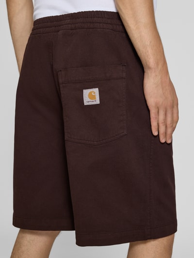 Mannenmodel draagt shorts uit katoenmix Model 'Floyde' van Carhartt Work In Progress in donkerbruin met achterzak en label, detailopname rugaanzicht. Context: Heren korte broek van Carhartt Work In Progress, mix van katoen en elastaan, relaxed fit, model 'Floyde', elastische band met tunnelkoord, zij-steekzakken, bandwijdte bij maat M: 39 cm, binnenbeenlengte bij maat M: 25 cm.