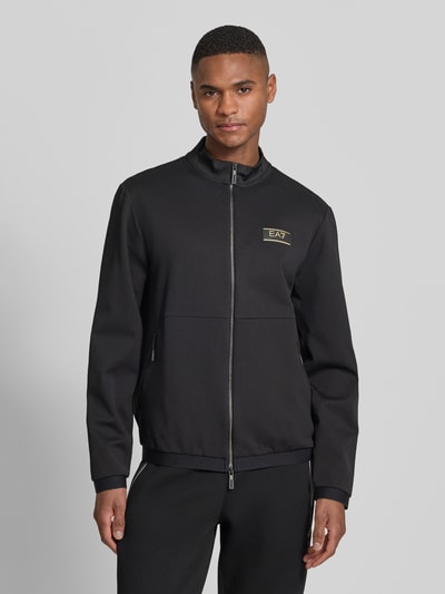 Męski model nosi Bluza rozpinana z zamkiem błyskawicznym dwustronnym od EA7 Emporio Armani, mieszanka materiałów z dodatkiem wiskozy, krój regular fit, stójka, naszywka z logo, kieszenie zapinane na zamek błyskawiczny, długość tyłu 65 cm, długość rękawów 65 cm, widok całego ciała z przodu.