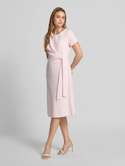 Frauenmodel trägt Knielanges Cocktailkleid mit Knotendetail und Reißverschluss von Rinascimento in zartem Rosa, Taillierter Schnitt, Rundhalsausschnitt, Knielänge, weiche Stofftextur, Rückseitiger Reißverschluss, Rückenlänge bei Größe S 105 cm, Brustweite bei Größe S 46 cm, Ärmellänge bei Größe S 17 cm, Ganzkörperansicht von vorne.