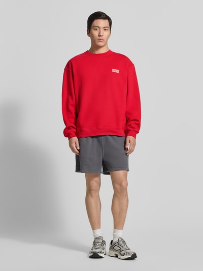 Męskamodel nosi czerwoną bluzę z logo, Reine Baumwolle, krój relaxed fit, okrągły dekolt, Sweatshirt mit Logo Modell 'PLIZZY' von American Vintage, długość tyłu 66 cm, długość rękawów 60 cm, Ganzkörperansicht von vorne