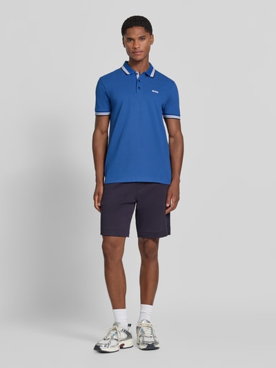 Männermodel trägt Regular Fit Poloshirt mit Label-Stitching Modell 'Paddy' von BOSS in Blau mit gerippten Abschlüssen, Ganzkörperansicht von vorne. Herren Poloshirt aus reiner Baumwolle, Polokragen, kurze Knopfleiste, Rückenlänge bei Größe M 73 cm.