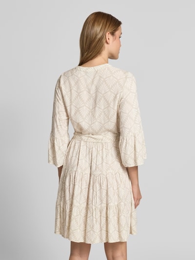 Frauenmodel trägt Tailliertes Tunikakleid aus reiner Viskose Modell 'MELBA' von Vero Moda, Optik:Allover-Muster, Trompetenärmel, Volant-Saum, Bindegürtel, Mini-Länge, Farbe Beige, fließendes Material, Ganzkörperansicht Rückansicht.