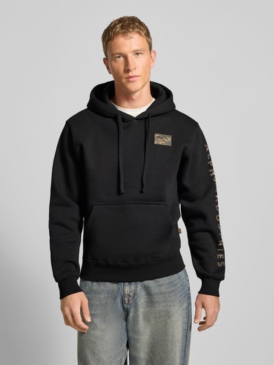Mannenmodel draagt Hoodie met Label-Stitching Model 'Camo Sleeve' van Alpha Industries in zwart katoenmix met kangoeroezak, regular fit, recht van voren, nahaufnahme oberkörper. Ruglengte maat M 67 cm, mouwlengte 66 cm.