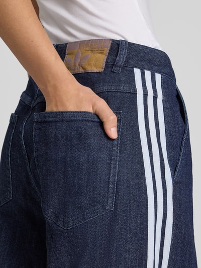 Vrouwenmodel draagt Loose Fit Jeans aus Baumwoll-Mix van adidas Originals, donkerblauw met witte strepen, detailopname achterzak.