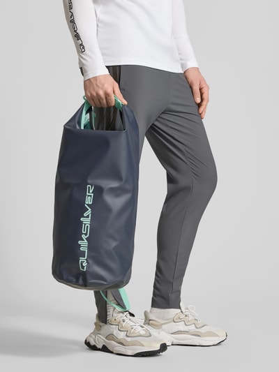 Männermodel trägt Tasche mit Riemen und Label-Print von Quiksilver in Grau mit grünem Akzent, Herren Tasche, Ganzkörperansicht von vorne.