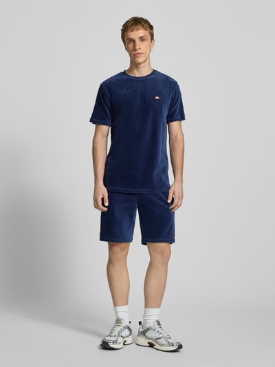 Męski model nosi T-Shirt mit Logo Modell 'MADORI' von Ellesse, mieszanka bawełny z dodatkiem streczu, krój regular fit, okrągły dekolt, logo, długość tyłu w rozmiarze M: 66 cm, ciemnoniebieski kolor, prążkowana faktura, całkowity widok z przodu.
