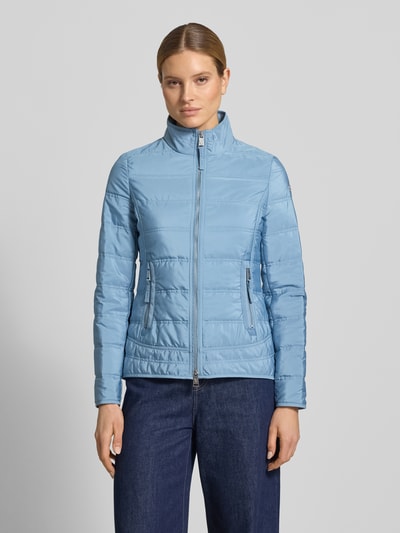 Kobiece modelka nosi kurtka pikowana z stójką i kieszeniami zapinanymi na zamek błyskawiczny 'Steppjacke mit Stehkragen und Reißverschlusstaschen' od Fuchs Schmitt, taliowany krój, jasnoniebieski kolor, detal z logo, długość tyłu w rozmiarze 36: 65 cm, szerokość ramion w rozmiarze 36: 45 cm, szerokość klatki piersiowej w rozmiarze 36: 42 cm, długość rękawów w rozmiarze 36: 65 cm, cała sylwetka z przodu.