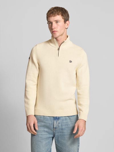 Männermodel trägt Slim Fit Pullover in Strick-Optik von Superdry, cremefarben, Stehkragen mit Reißverschluss, Logo-Stitching, Ganzkörperansicht von vorne.