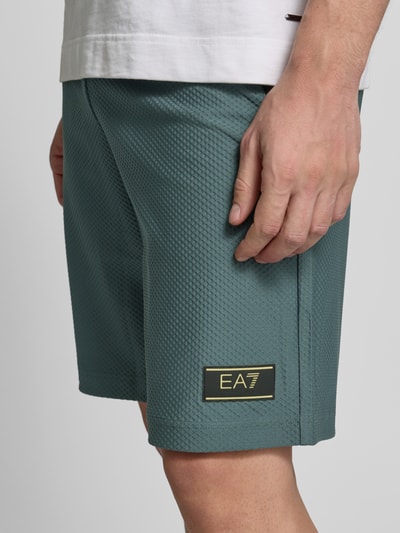 Mężczyznę model ma na sobie Regular Fit Shorts mit Strukturmuster od EA7 Emporio Armani, elastyczna mieszanka materiałów, krój regular fit, wzór strukturalny, elastyczny pas ze sznurkiem, szerokość w rozmiarze M: 41 cm, długość wewnętrzna nogawki w rozmiarze M: 26 cm, zbliżenie na dolną część ciała.