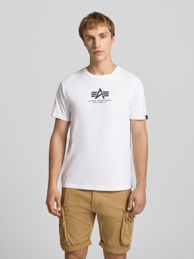 Männermodel trägt T-Shirt mit Logo und Rundhalsausschnitt van Alpha Industries, witte kleur, regular fit van katoen, logo op borst, Ganzkörperansicht von vorne.