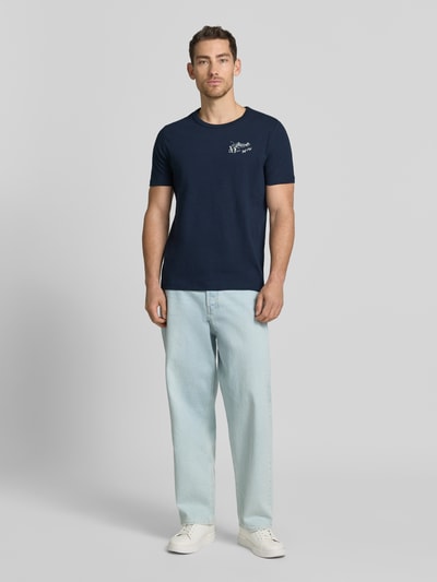 Męski model nosi Regular Fit T-Shirt aus reiner Baumwolle od s.Oliver RED LABEL, efekt: jednolity kolor, krój regular fit, nadruk z motywem, rękaw: krótki rękaw, materiał: bawełna, ciemnoniebieski, jasnoniebieskie spodnie, białe buty, całopostaciowy widok z przodu.