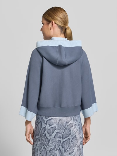 Frauenmodel trägt Relaxed Fit Sweatjacke aus Viskose-Mix mit Logo-Detail von Marc Cain, Blau- und Grautöne, lockerer Schnitt, Rückenansicht, Kontext: Damen Sweatjacke, Viskose-Mix, Kapuze mit Tunnelzug, Reißverschluss, elastischer Bund, Rückenlänge 51 cm, Ärmellänge 54 cm.