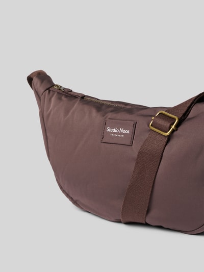 Produktfoto, Fanny Pack mit Logo-Detail Modell 'Puffy' von Studio Noos, glatter brauner Stoff, goldfarbene Metallelemente, Kontext: Damen Fanny Pack, Hauptfach mit Reißverschluss, verstellbarer Schulterriemen, Maße: 41x20x10 cm, Schulterriemen 108 cm.