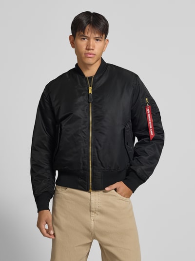 Männermodel trägt Bomberjacke mit Pattentaschen und Label-Applikation von Alpha Industries in Schwarz, Reißverschluss, Nahaufnahme Oberkörper. Regular Fit, Stehkragen, rote Label-Applikation am Ärmel, glatte Oberfläche, sichtbare Strukturdetails, Kontext: Rückenlänge 69 cm, Schulterbreite 44 cm, Brustweite 54 cm, Ärmellänge 64 cm.