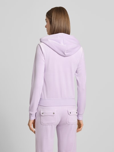 Frauenmodel trägt Slim Fit Sweatjacke aus Samt Modell 'Robertson' von Juicy Couture in Lila mit Kapuze, Tunnelzug, Känguru-Tasche, Ganzkörperansicht Rückansicht.