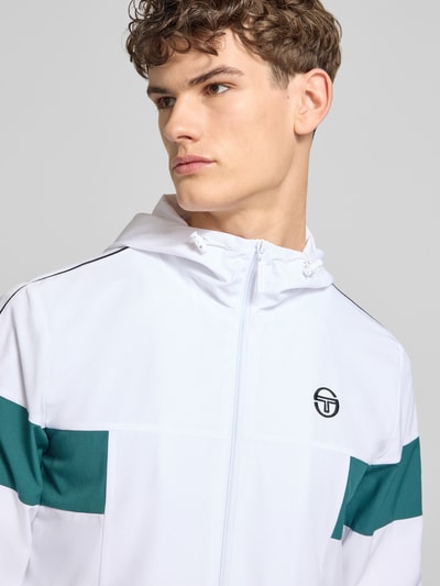Männermodel trägt Regular Fit Jacke mit Logo-Stitching Modell 'Caspio' von SERGIO TACCHINI in Weiß-Grün, Nahaufnahme Oberkörper. Elastischer Material-Mix, Kapuze mit Tunnelzug, Reißverschluss und Eingrifftaschen integriert.