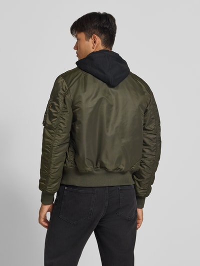 Männermodel trägt Jacke mit Reißverschlusstasche am Ärmel von Alpha Industries in Olivgrün, glatte Struktur, gerippte Abschlüsse, Ganzkörperansicht Rückansicht. Die Jacke hat Kapuze mit Reißverschluss, Label-Patch und Eingrifftaschen mit Druckknöpfen, Regular Fit.