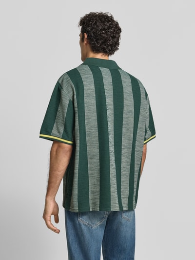 Männermodel trägt Loose Fit Poloshirt mit V-Ausschnitt von Les Deux in Grün-Grau mit Strukturmuster, Kontrastkante, Ganzkörperansicht Rückansicht. Material-Mix mit Viskose-Anteil, Label-Stitchings, Rückenlänge bei Größe M: 74 cm.