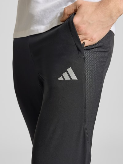Männermodel trägt Regular Fit Trackpants mit elastischem Logo-Bund von adidas Training, unifarben, seitliche Reißverschlusstasche, Nahaufnahme Oberschenkel.